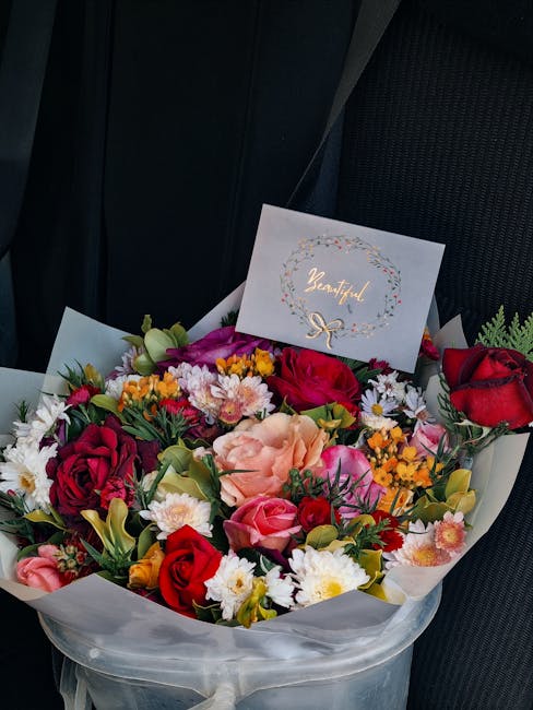 A vibrant bouquet of fresh flowers arranged with roses in red, pink, and peach shades, complemented by yellow, white, and purple blooms, including daisies and chrysanthemums. The bouquet is wrapped in
